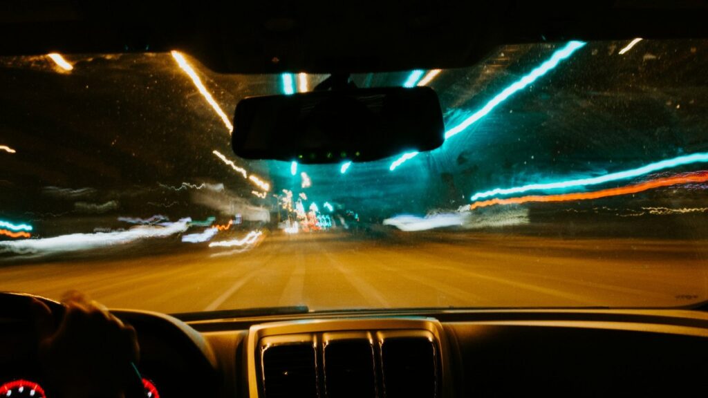 夜の街を走る車の運転席からの視点。ハンドルを握る手とフロントガラス越しに流れる街灯りが写っている