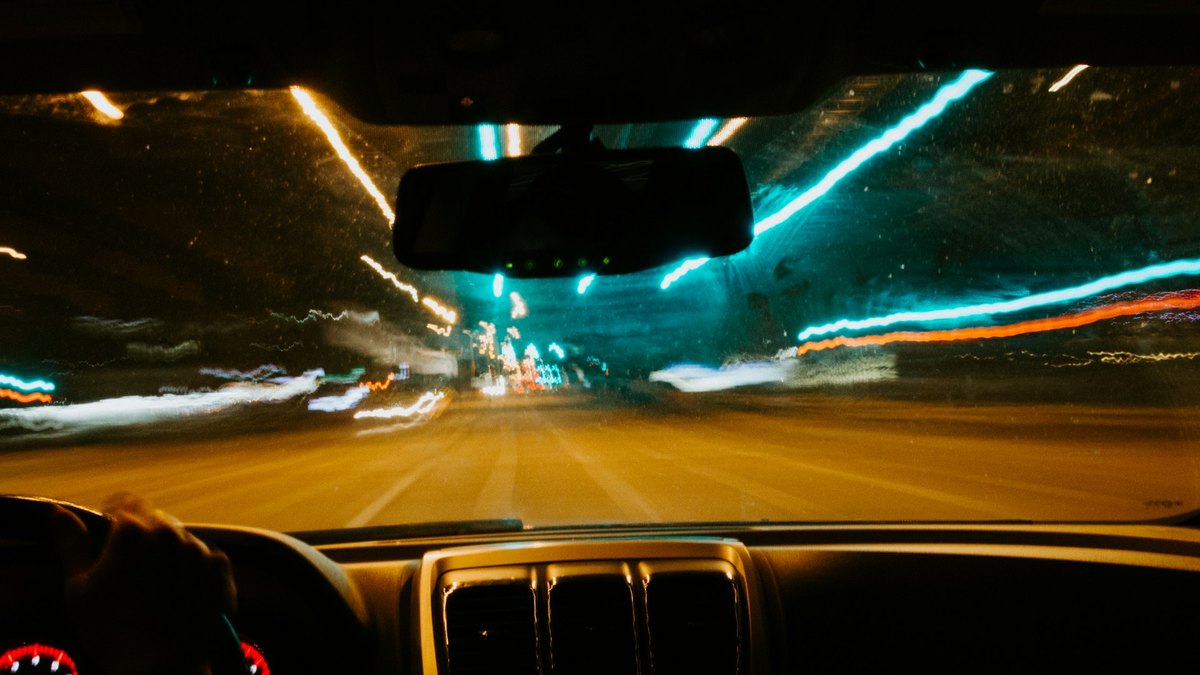 夜の街を走る車の運転席からの視点。ハンドルを握る手とフロントガラス越しに流れる街灯りが写っている
