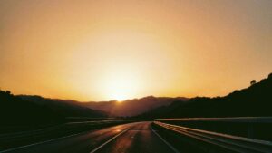 夜明けの高速道路。ガードレールの先に朝日が昇り、路面がオレンジ色に光っている